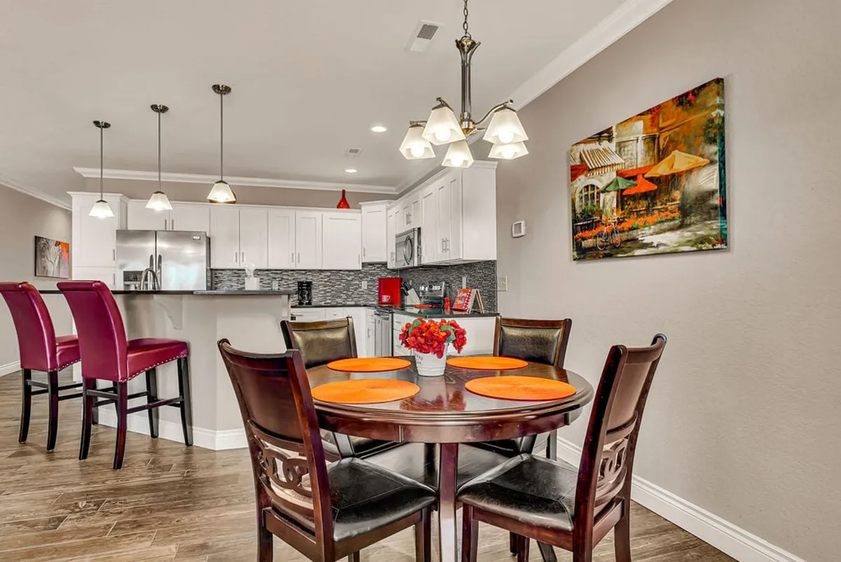 A warm, inviting dining area featuring a round dining table set with orange plates, bar stools lining the kitchen island, visible kitchen appliances, and tasteful wall art. The setup is ready for family dinners, casual gatherings, and entertaining guests.