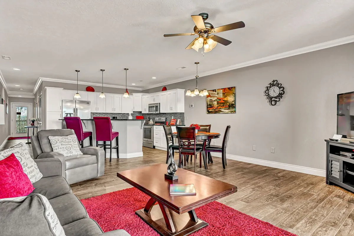 This image captures an inviting open-concept living and dining area. A sofa with colorful cushions is set beside a coffee table, with the dining table and kitchen island visible in the background. Large windows provide ample natural light, suggesting views of the outdoors. The layout exemplifies a space designed for both relaxation and group gatherings.