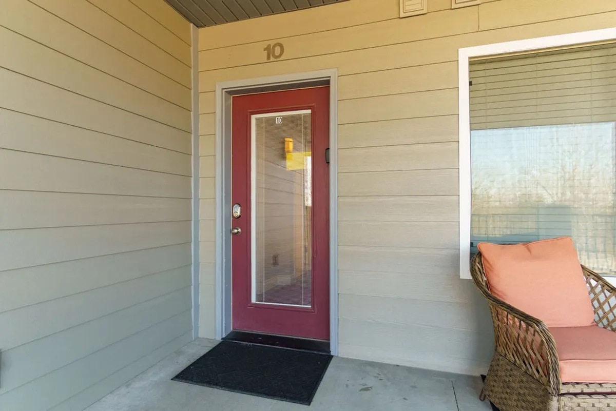 A warm exterior shot showing the condo's inviting red front door, a wicker chair on a small porch, and the number 10 on the door. The porch scene suggests a welcoming entry, perfect for group arrivals or enjoying quiet moments before heading out for nearby adventures.
