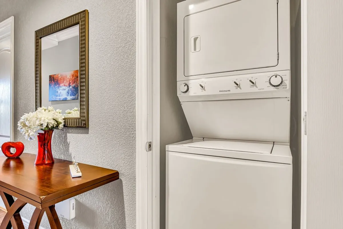 A functional yet stylish in-unit laundry area showcasing a stacked washer and dryer, a small wooden table, decorative elements like a floral arrangement, and a framed mirror. The space is clean and practical, offering convenience for longer stays.