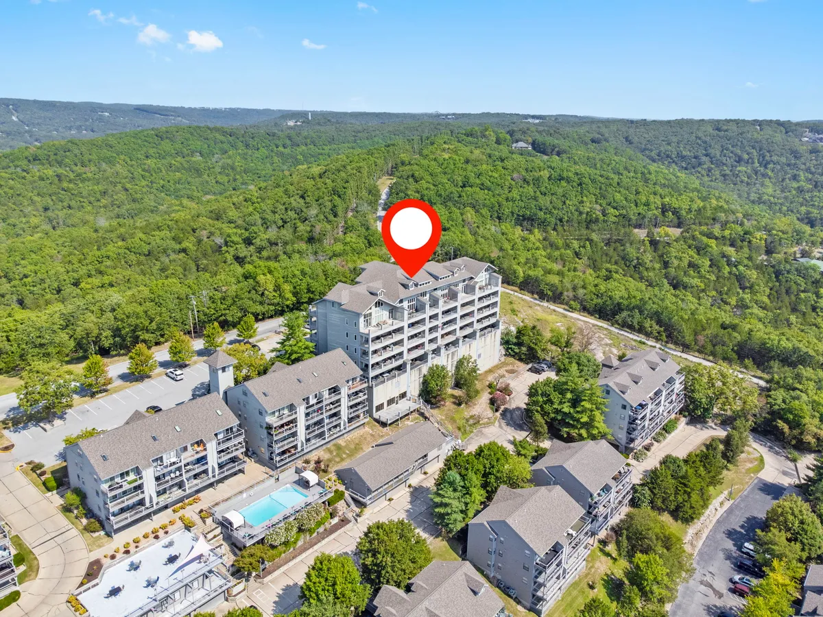 A broad exterior shot showing the main building nestled in greenery, a sparkling swimming pool in the foreground, and wooded walking trails winding nearby. The setting is bright and inviting, emphasizing outdoor recreation and the natural beauty of the area.